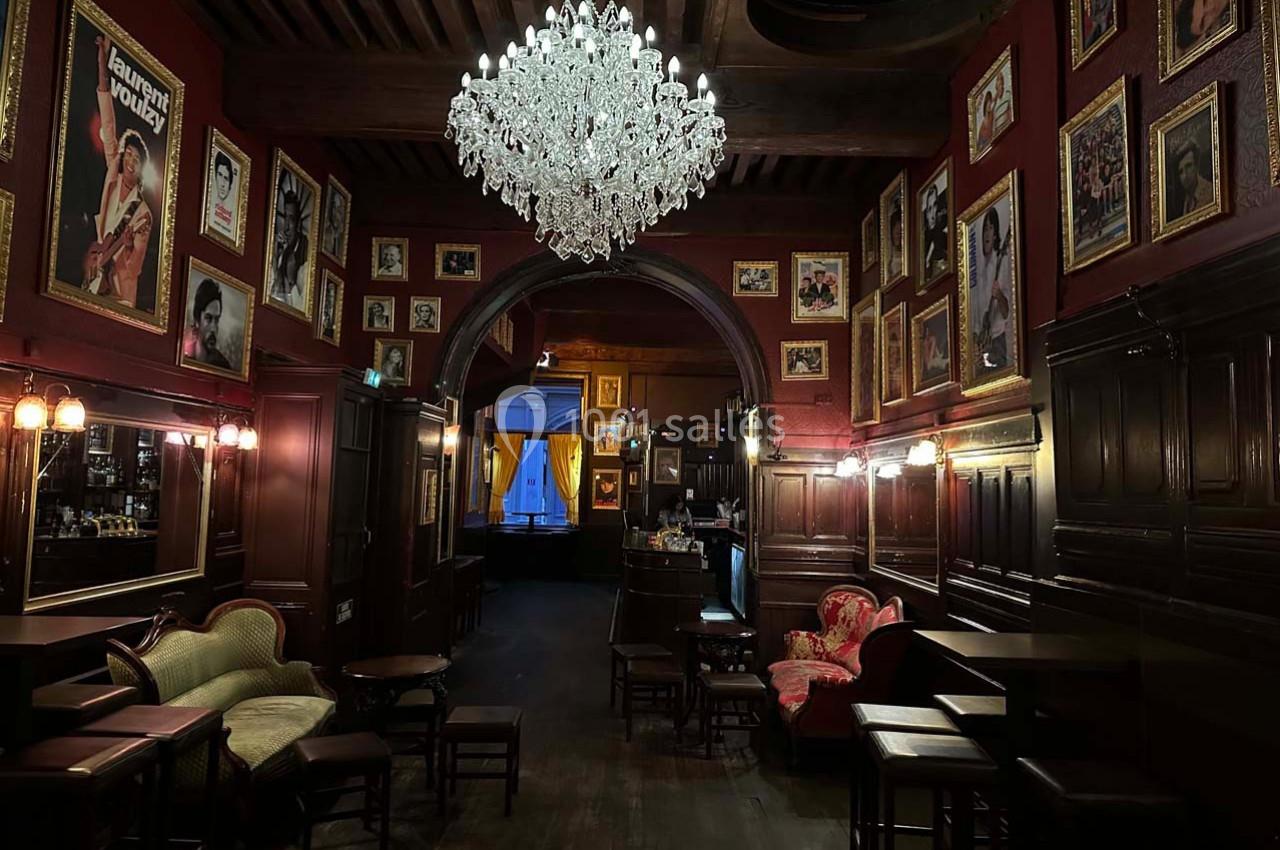 Salle de bar au décor vintage avec murs rouges ornés de cadres, lustre en cristal et mobilier ancien.