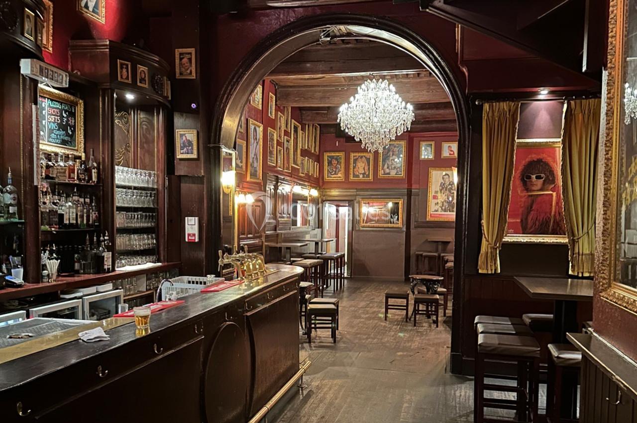 Intérieur d'un bar chaleureux avec comptoir en bois, lustre central, murs rouges ornés de cadres et tables disposées.