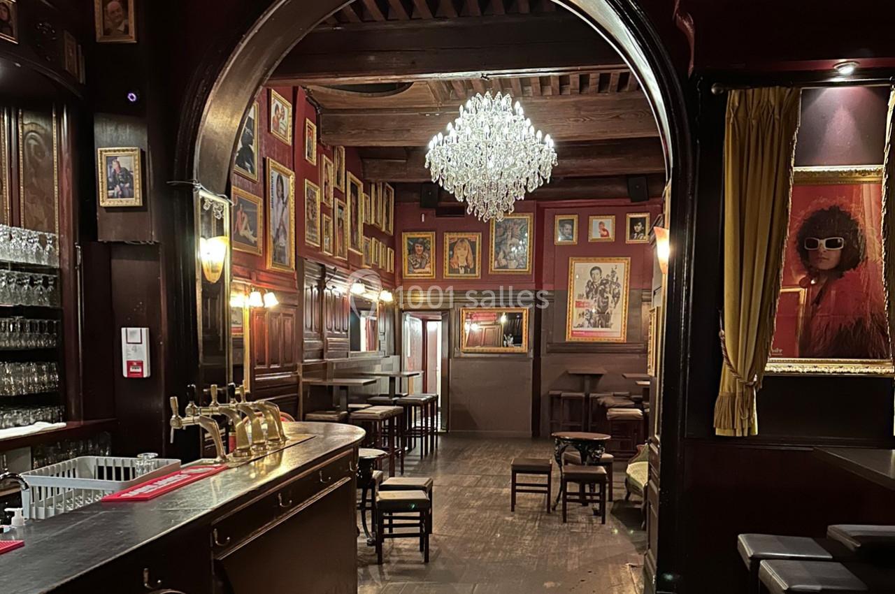 Intérieur d'un bar avec un lustre central, des murs ornés de cadres et un comptoir en bois sombre.