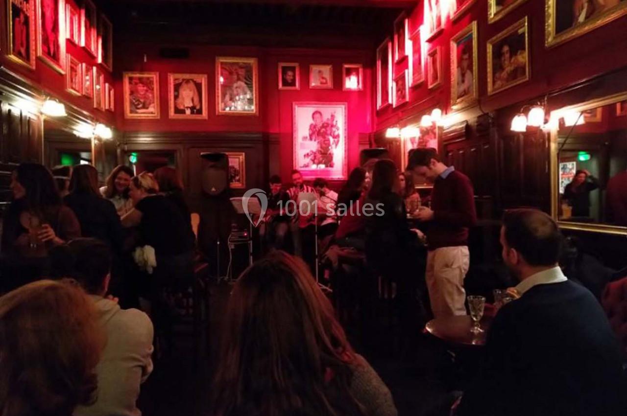 Salle de bar chaleureuse avec éclairage rouge, murs ornés de cadres, et musiciens jouant devant un public.