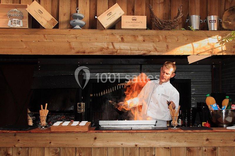 Chef cuisinier en train de flamber un plat dans une cuisine ouverte en bois, avec des accessoires culinaires autour.