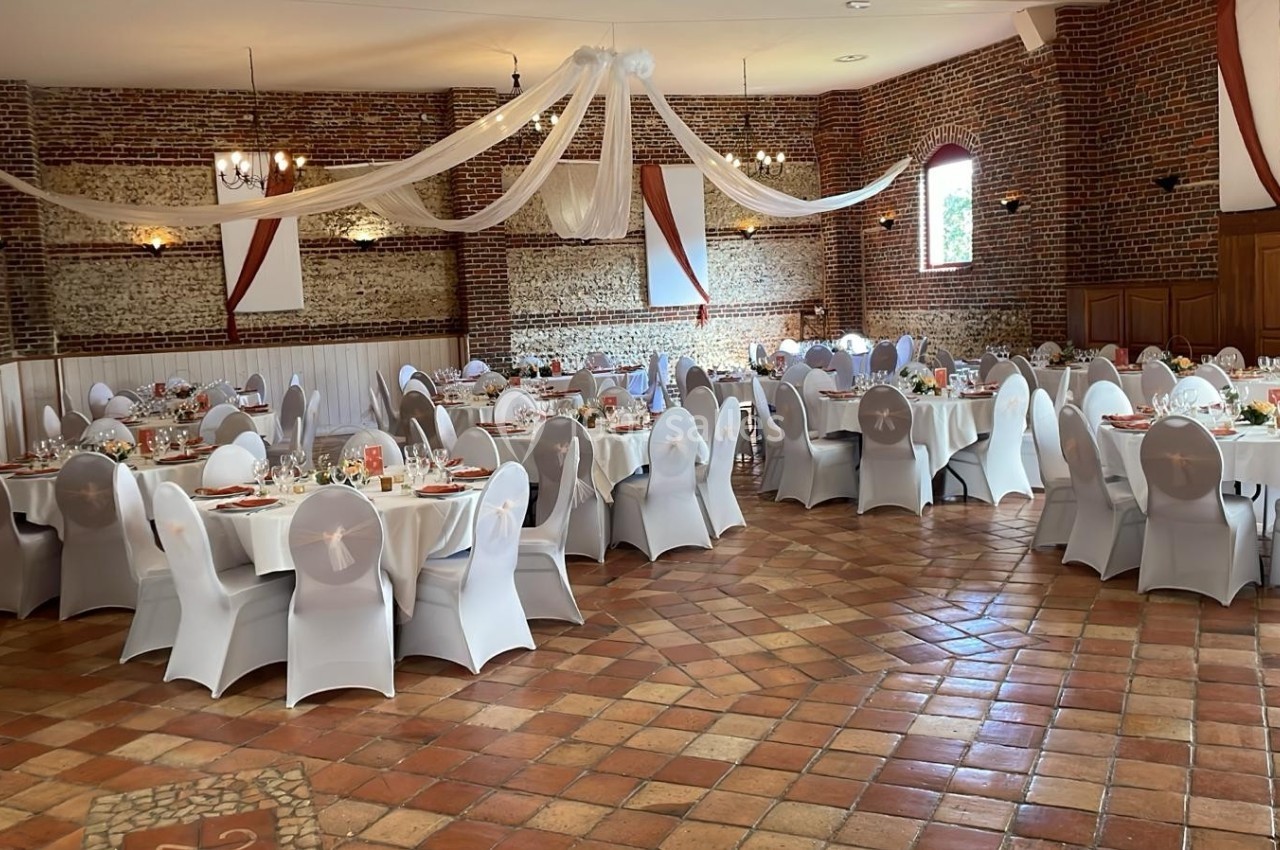 Salle de réception décorée avec des tables rondes dressées, chaises blanches et voilages suspendus au plafond.
