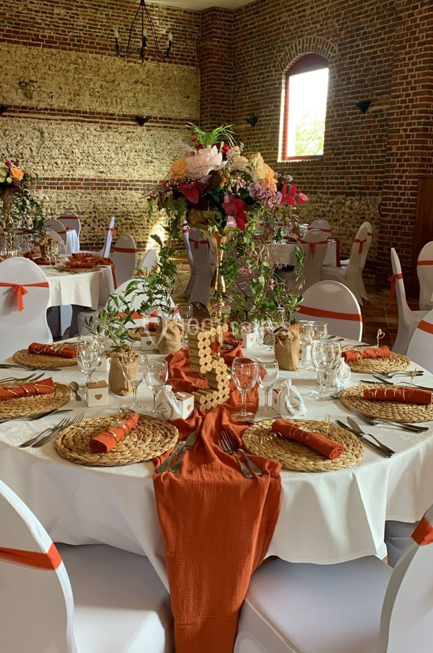 Table de réception décorée avec des nappes blanches, chemins de table orange, fleurs et vaisselle élégante dans une salle en…