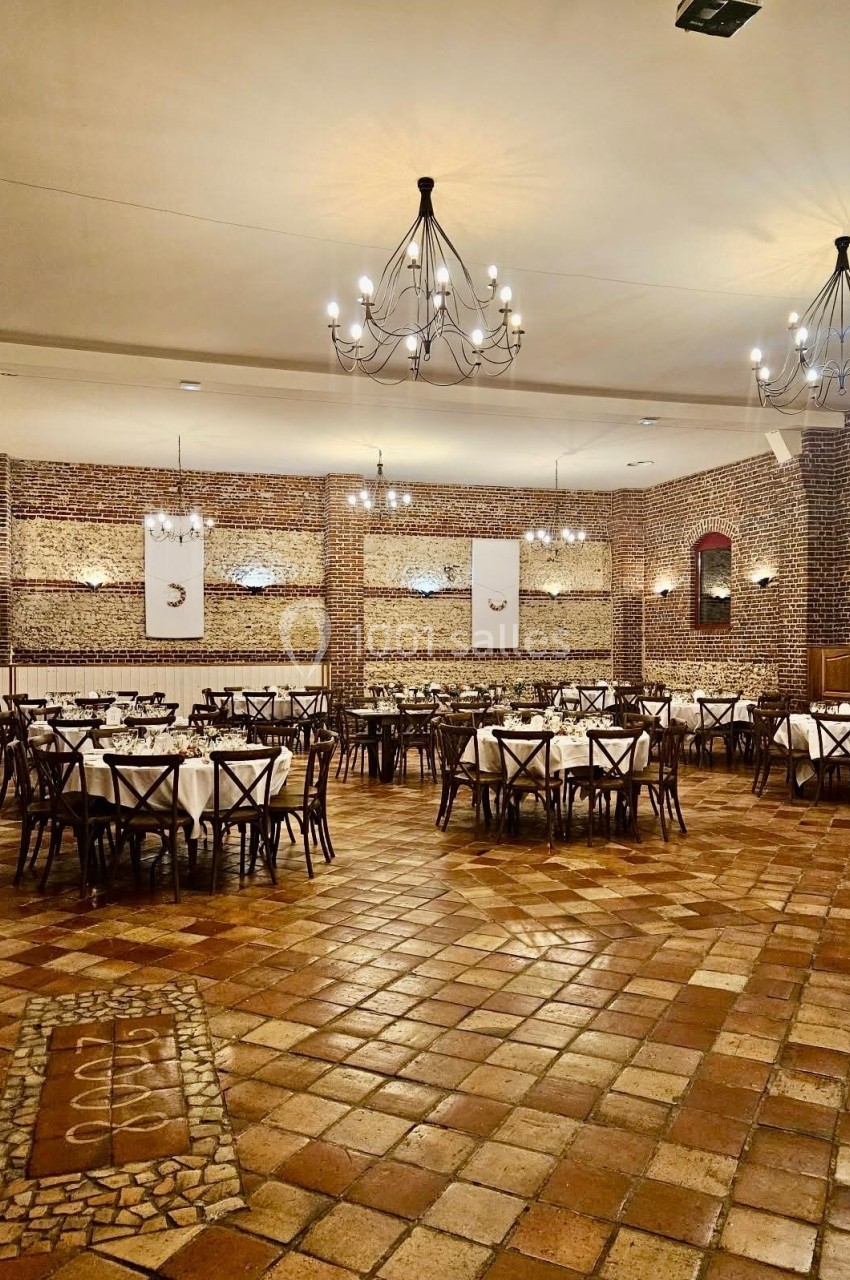 Salle de réception avec murs en briques, tables rondes dressées et lustres suspendus au plafond.
