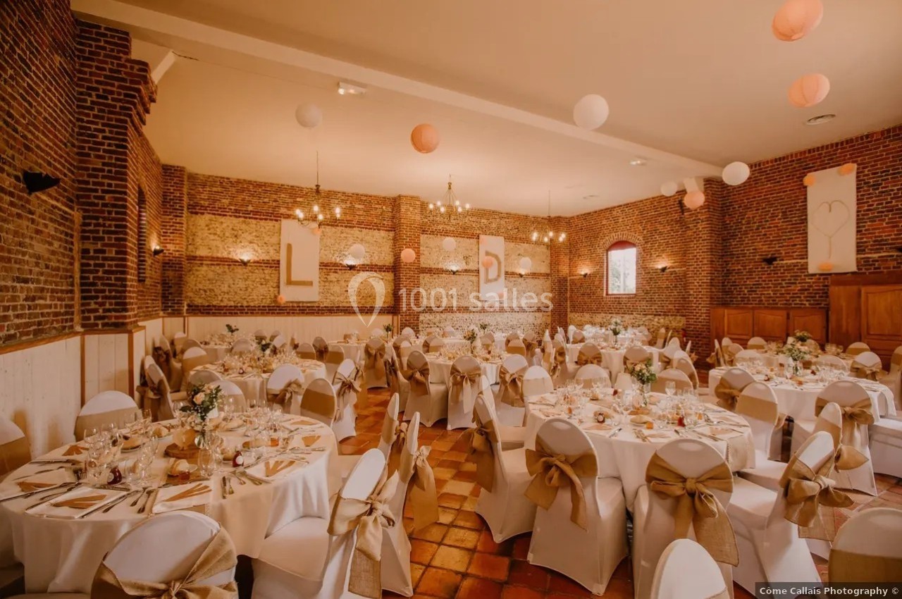 Salle de réception décorée avec des tables rondes, nappes blanches, chaises ornées de nœuds dorés et éclairage chaleureux.