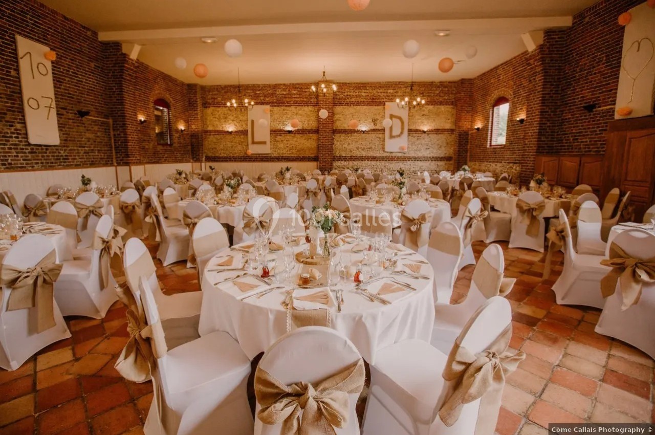 Salle de réception décorée pour un mariage, avec tables rondes dressées, chaises blanches et murs en briques apparentes.