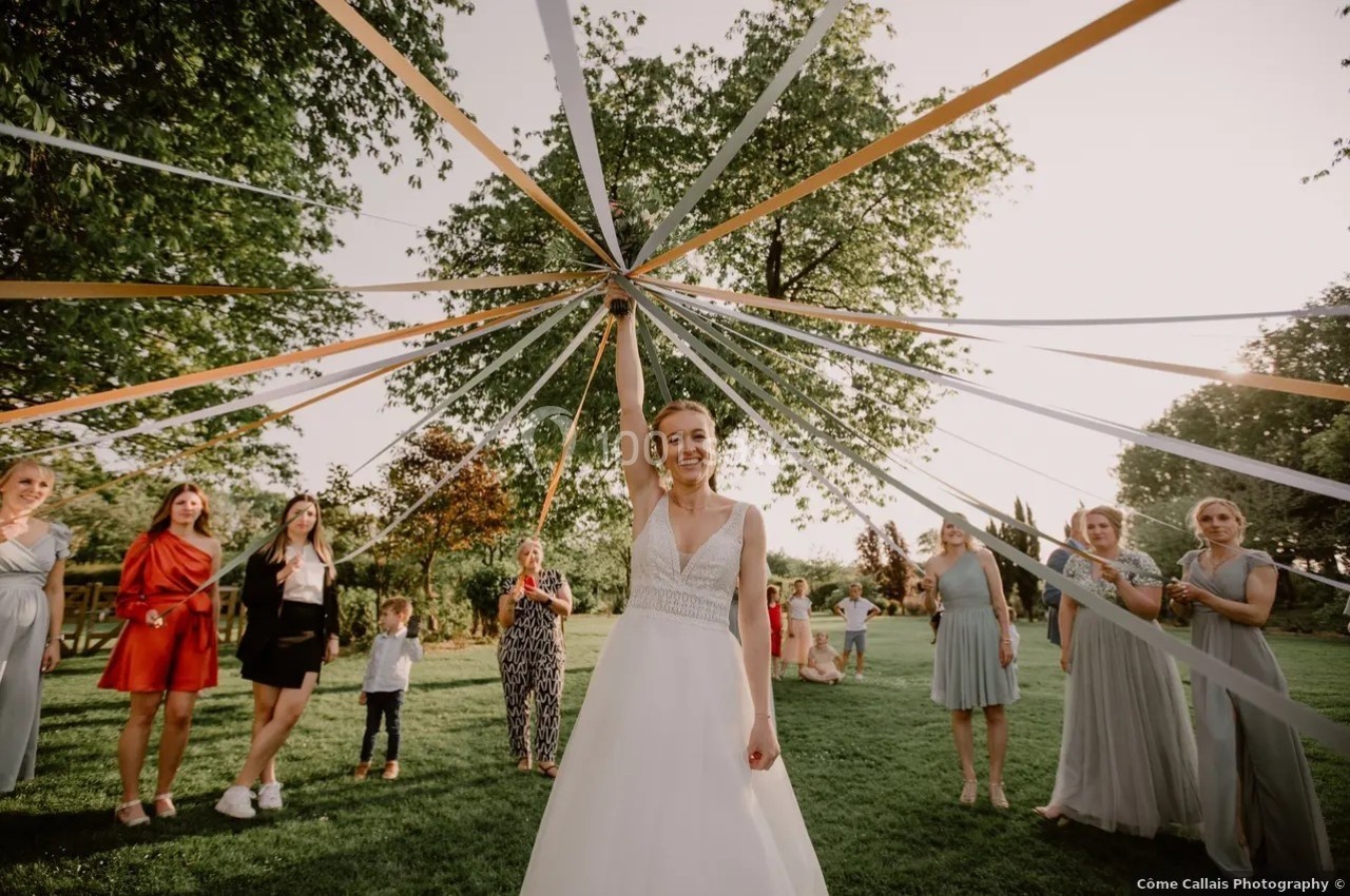 Une mariée souriante tient des rubans tendus en cercle, entourée d'invités dans un jardin ensoleillé.
