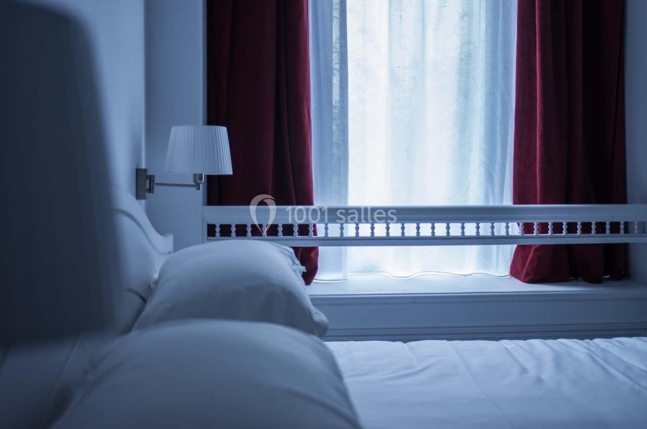 Lit avec draps blancs près d'une fenêtre encadrée de rideaux rouges, éclairée par une lampe de chevet.