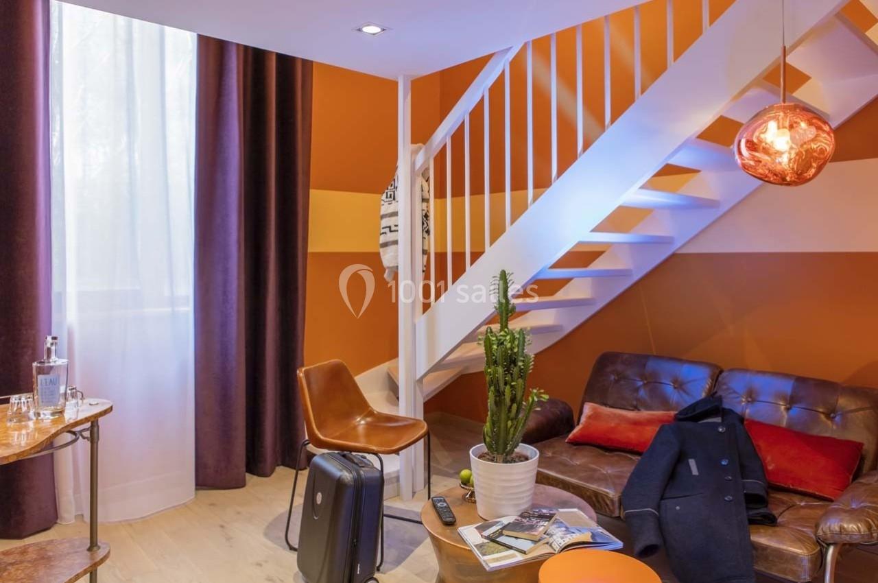 Salon moderne avec escalier blanc, murs orange, canapé en cuir, valise au sol et plante en pot.