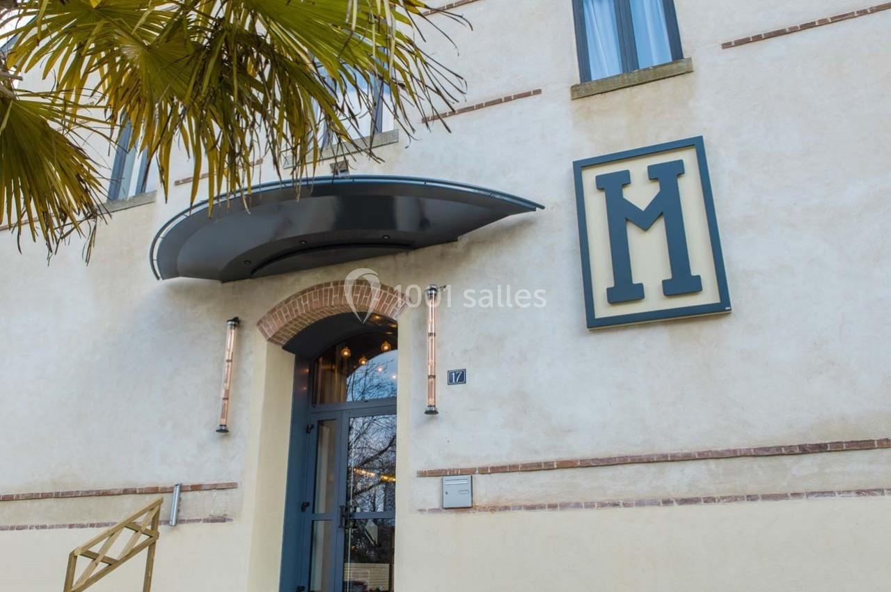 Façade d'un bâtiment beige avec une porte vitrée, un auvent métallique et un grand logo en forme de ’M’.