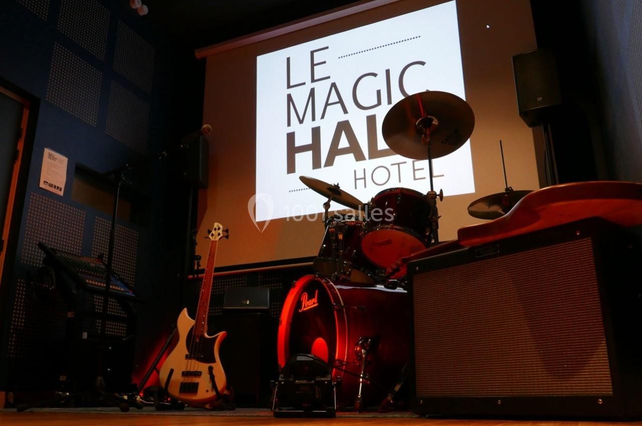 Scène de musique avec batterie, guitare basse, amplificateur et écran affichant ’Le Magic Hall Hotel’.