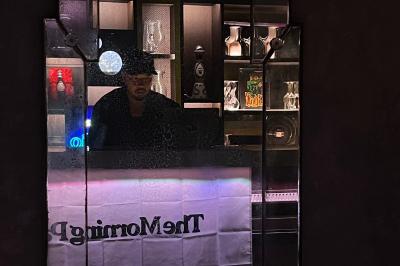 Un homme portant un chapeau est derrière un comptoir, reflété dans un miroir entouré d'étagères éclairées.