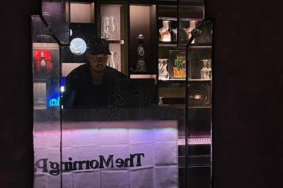 Un homme derrière un comptoir éclairé, entouré de bouteilles et d'objets décoratifs dans un bar sombre.