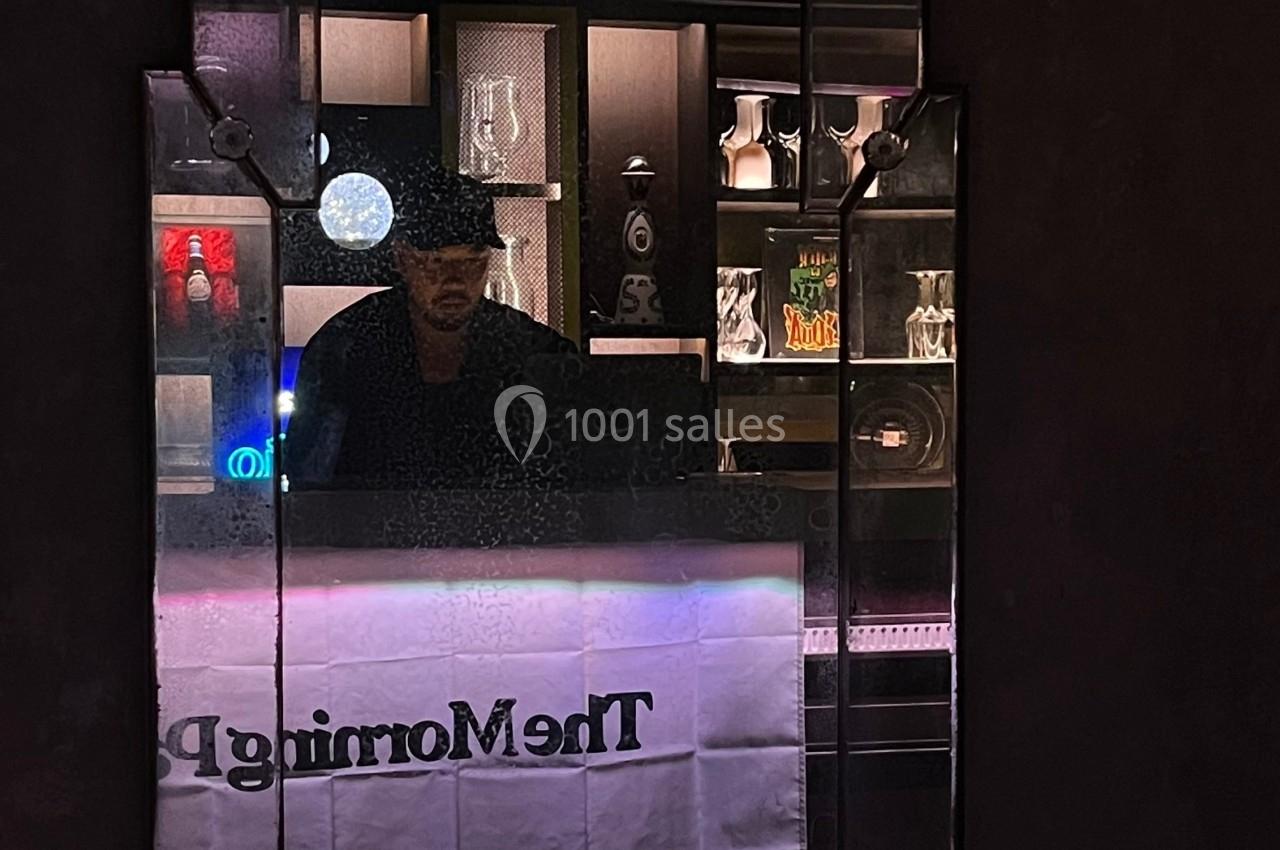 Un homme derrière un comptoir éclairé, entouré de bouteilles et d'objets décoratifs dans un bar sombre.