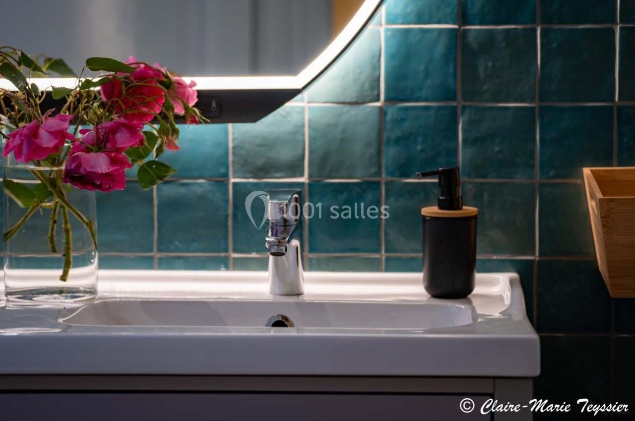 Lavabo avec robinet chromé, distributeur de savon noir et bouquet de fleurs roses devant un mur de carreaux verts.