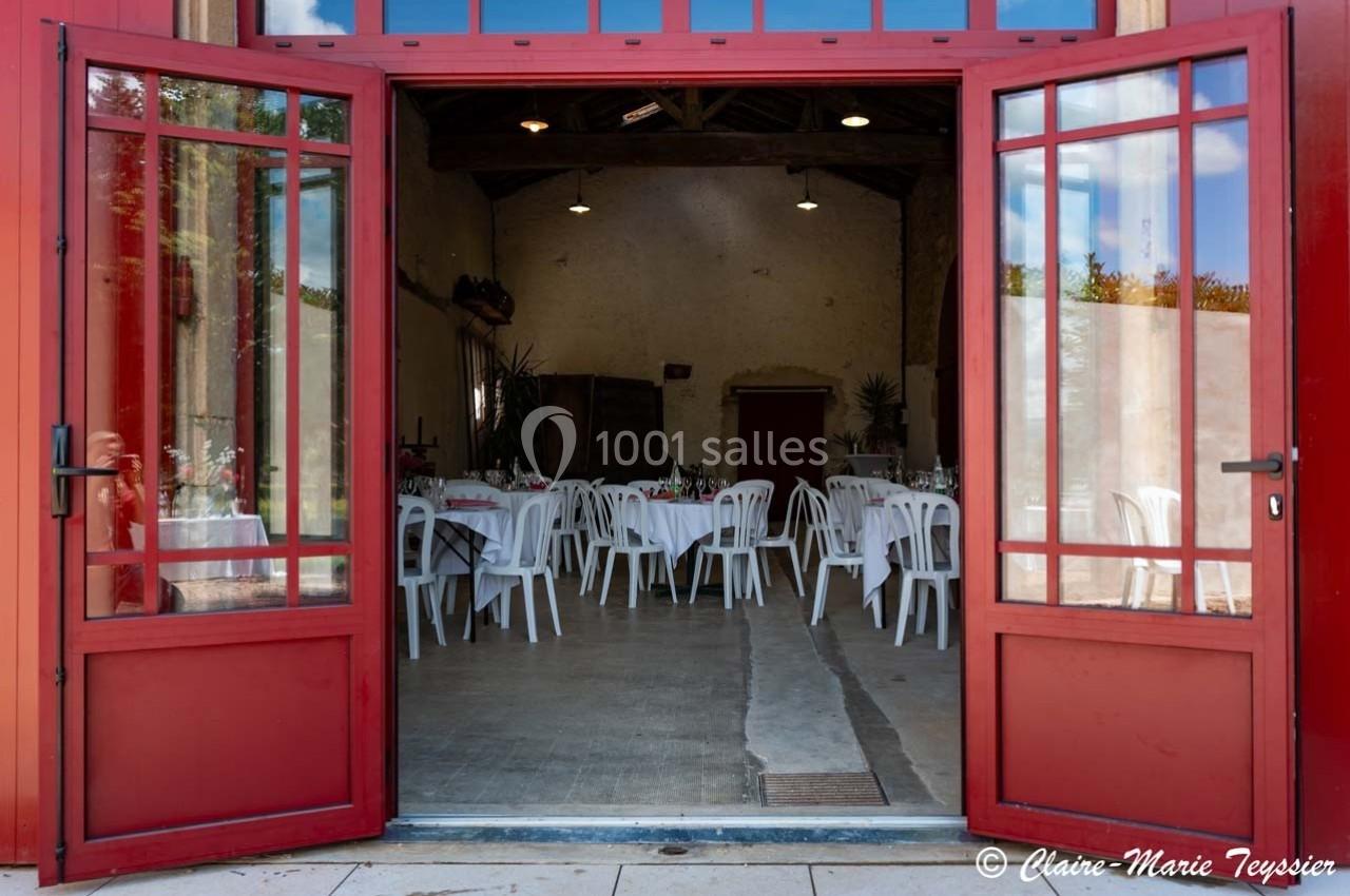 Salle de réception avec des tables dressées et des chaises blanches, vue à travers des portes vitrées rouges ouvertes.