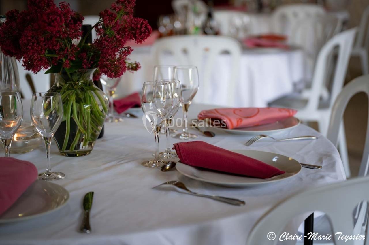 Table dressée avec nappes blanches, serviettes roses, verres et couverts, décorée d'un bouquet de fleurs rouges.