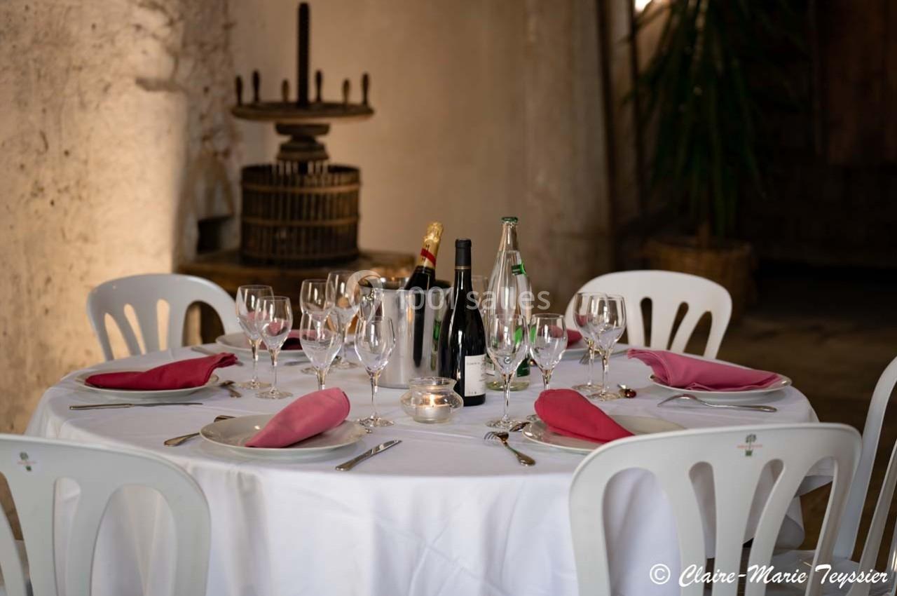 Table ronde dressée avec nappes blanches, serviettes roses, verres, bouteilles et bougie dans un cadre rustique.