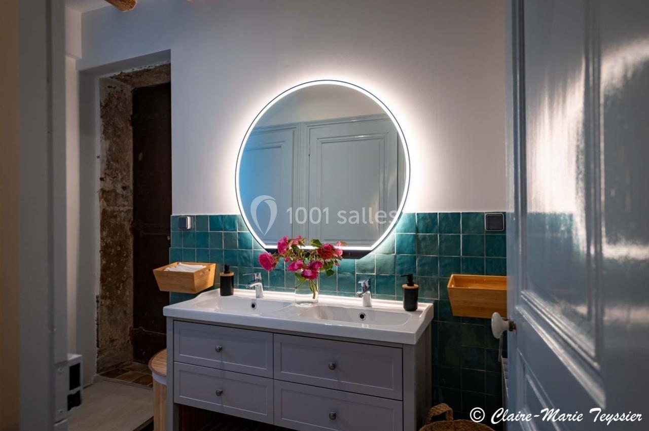 Salle de bain avec un miroir rond éclairé, un plan vasque double blanc et des carreaux muraux verts.