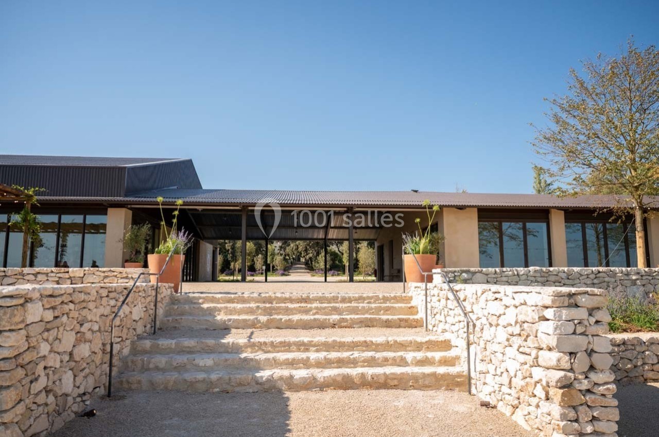 Entrée d'un bâtiment moderne avec toit en métal, entourée de murs en pierre et de plantes en pots sous un ciel dégagé.