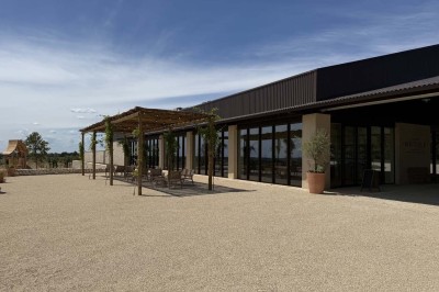 Terrasse ombragée avec pergola en bois, tables et chaises devant un bâtiment moderne avec de grandes baies vitrées.