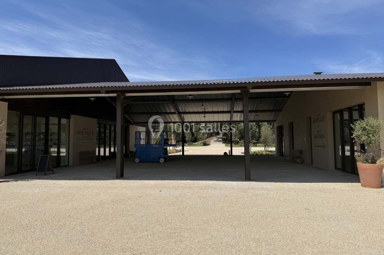 Structure couverte avec toit en tôle, entourée de bâtiments beige et d'une cour en gravier sous un ciel dégagé.