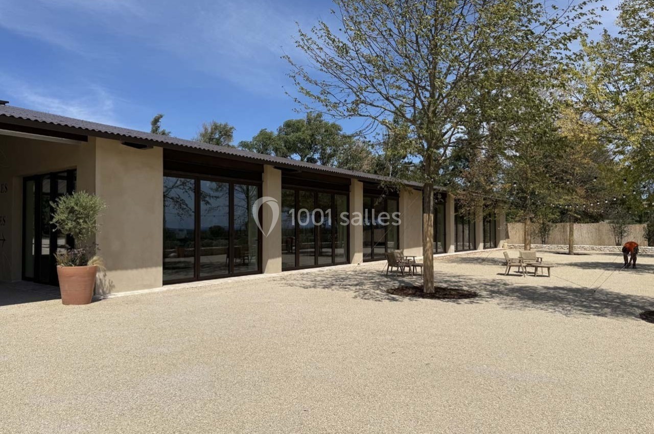 Bâtiment moderne avec de grandes baies vitrées, entouré d'arbres et d'une cour en gravier sous un ciel dégagé.