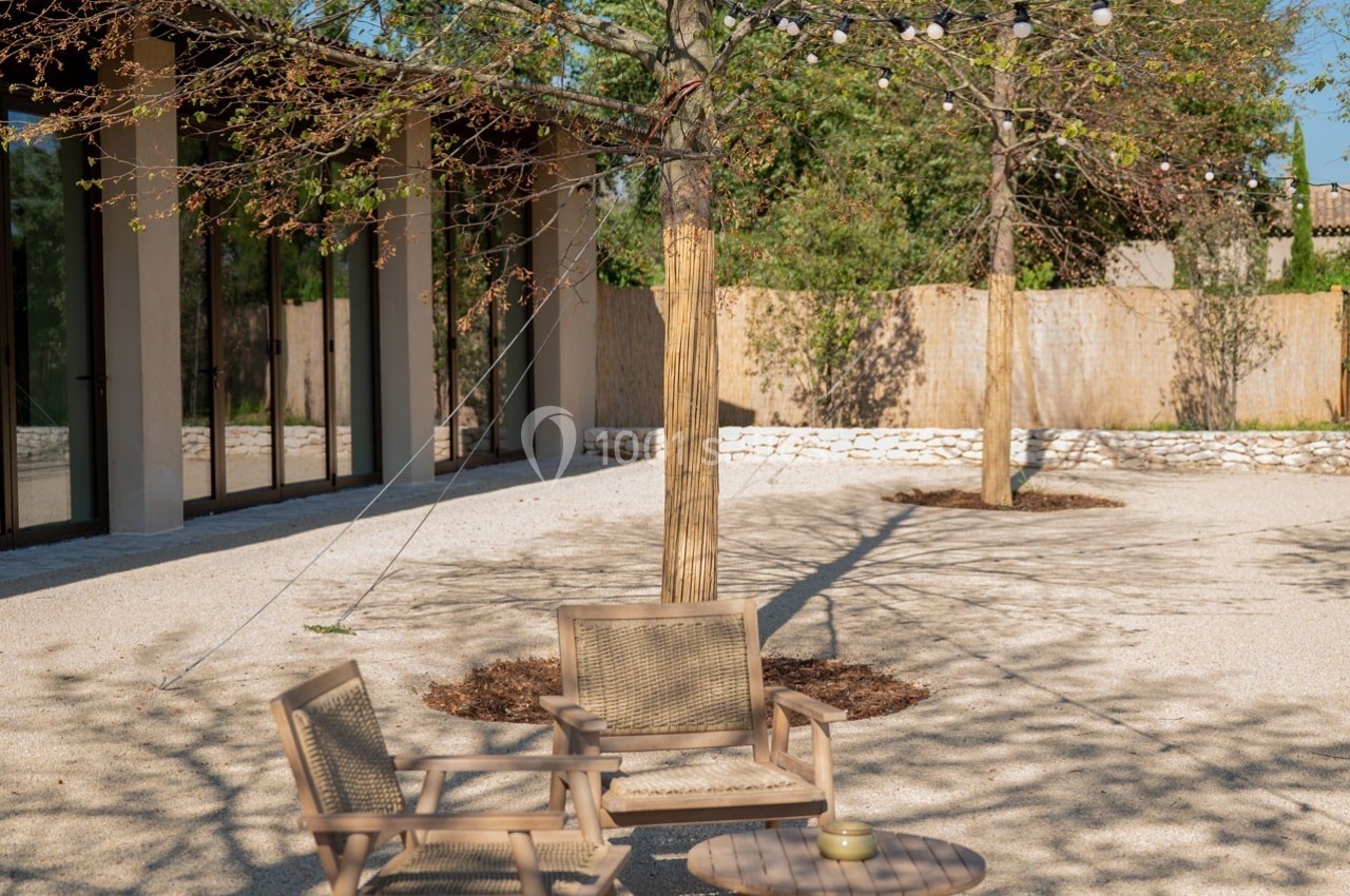 Mobilier de jardin en bois sous des arbres avec guirlandes lumineuses, dans une cour ensoleillée et gravillonnée.
