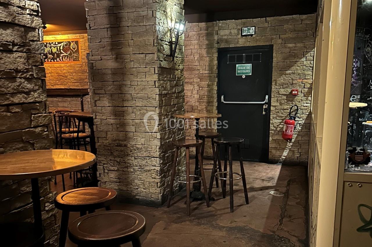 Intérieur d'un bar avec murs en briques, tables hautes en bois, et une porte de secours au fond.