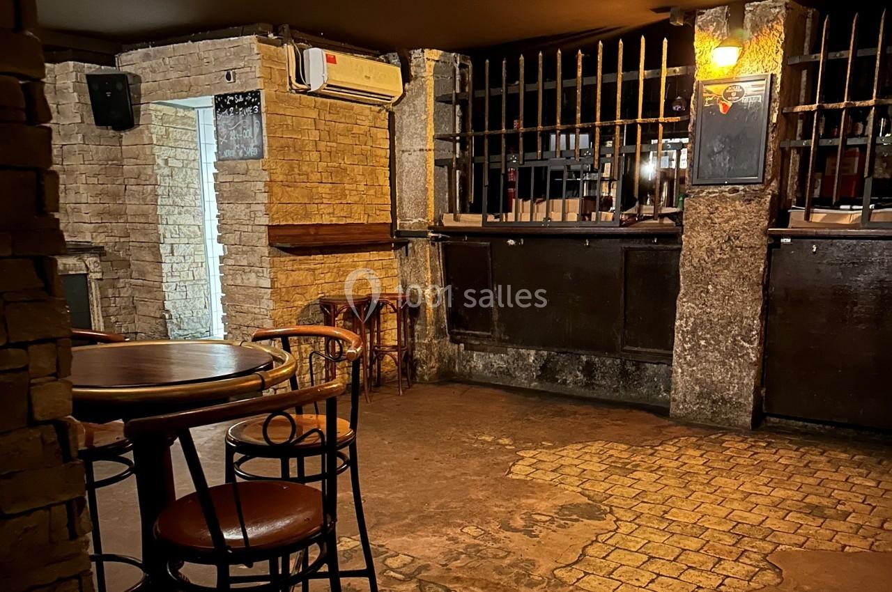 Salle intérieure avec murs en pierre, tables et chaises en bois, éclairage tamisé et comptoir avec grille métallique.