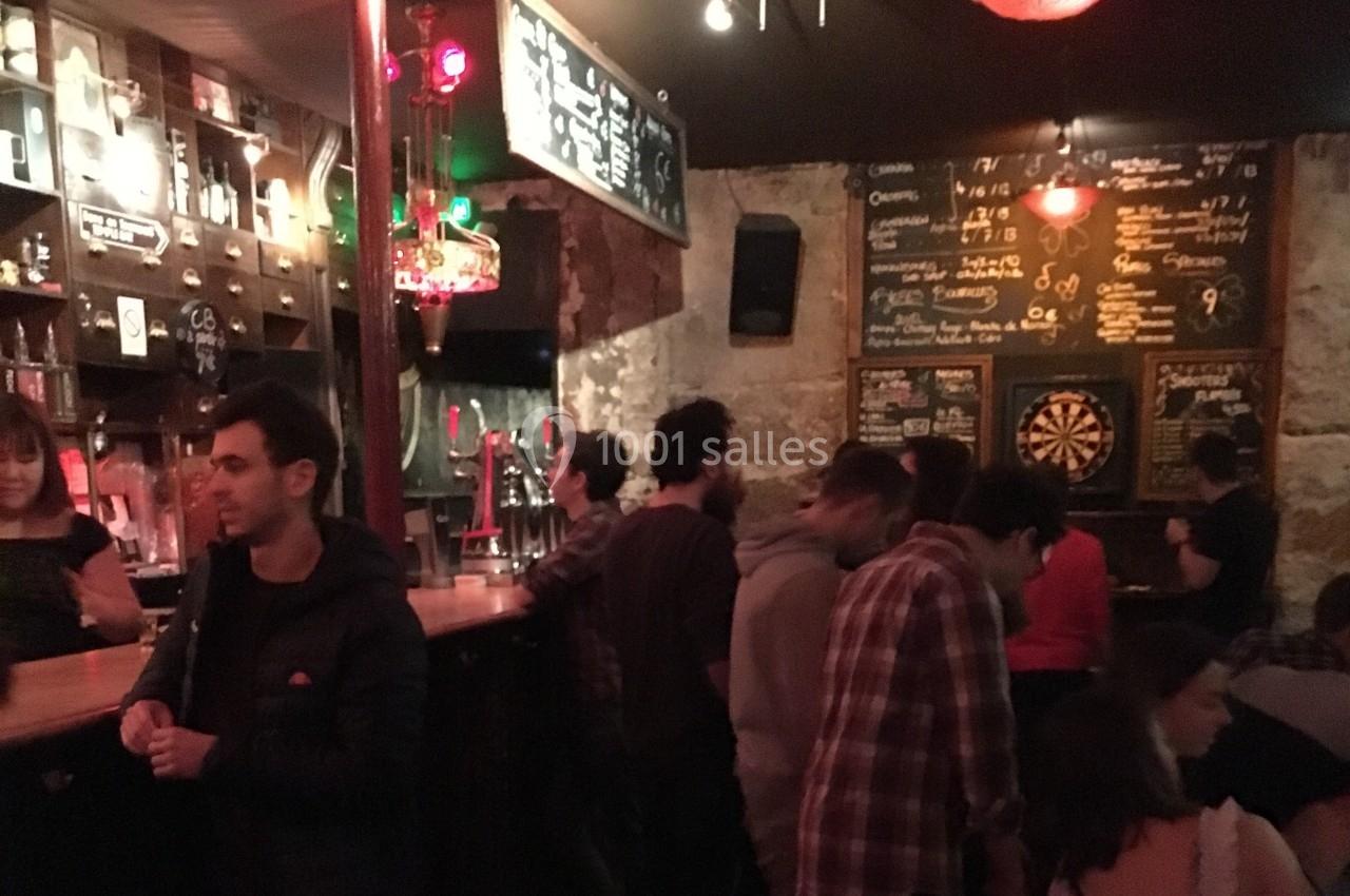 Des personnes discutent dans un bar à l'ambiance tamisée, avec un tableau de fléchettes et des menus écrits à la craie.
