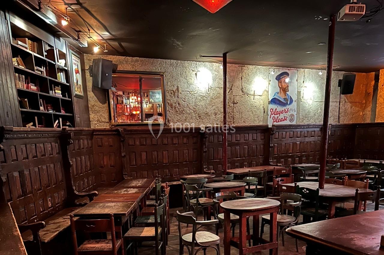 Salle de bar rustique avec tables en bois, étagères de livres, éclairage tamisé et affiche murale décorative.