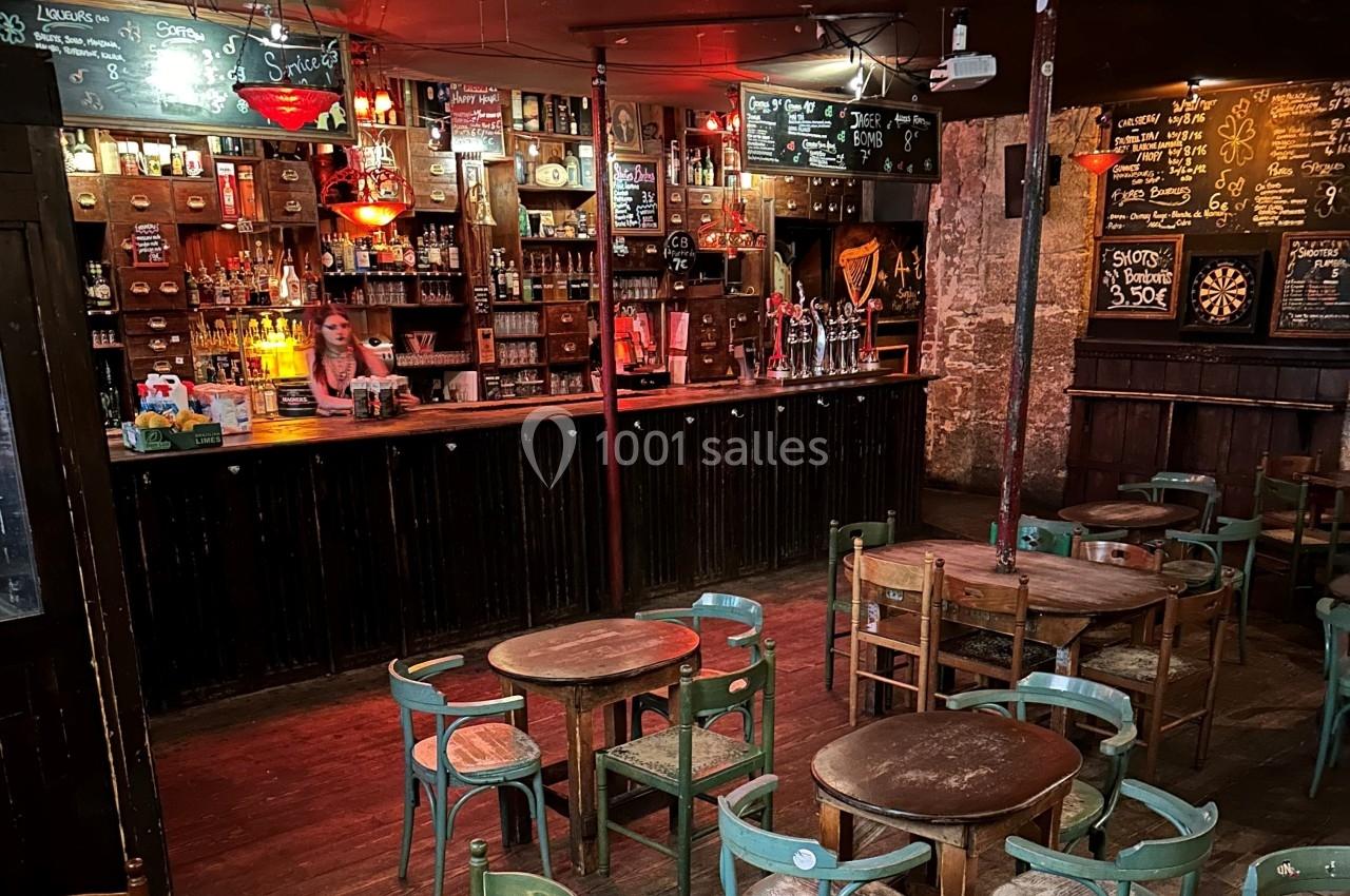 Intérieur d'un bar rustique avec tables en bois, chaises dépareillées, comptoir éclairé et ardoises affichant les menus.