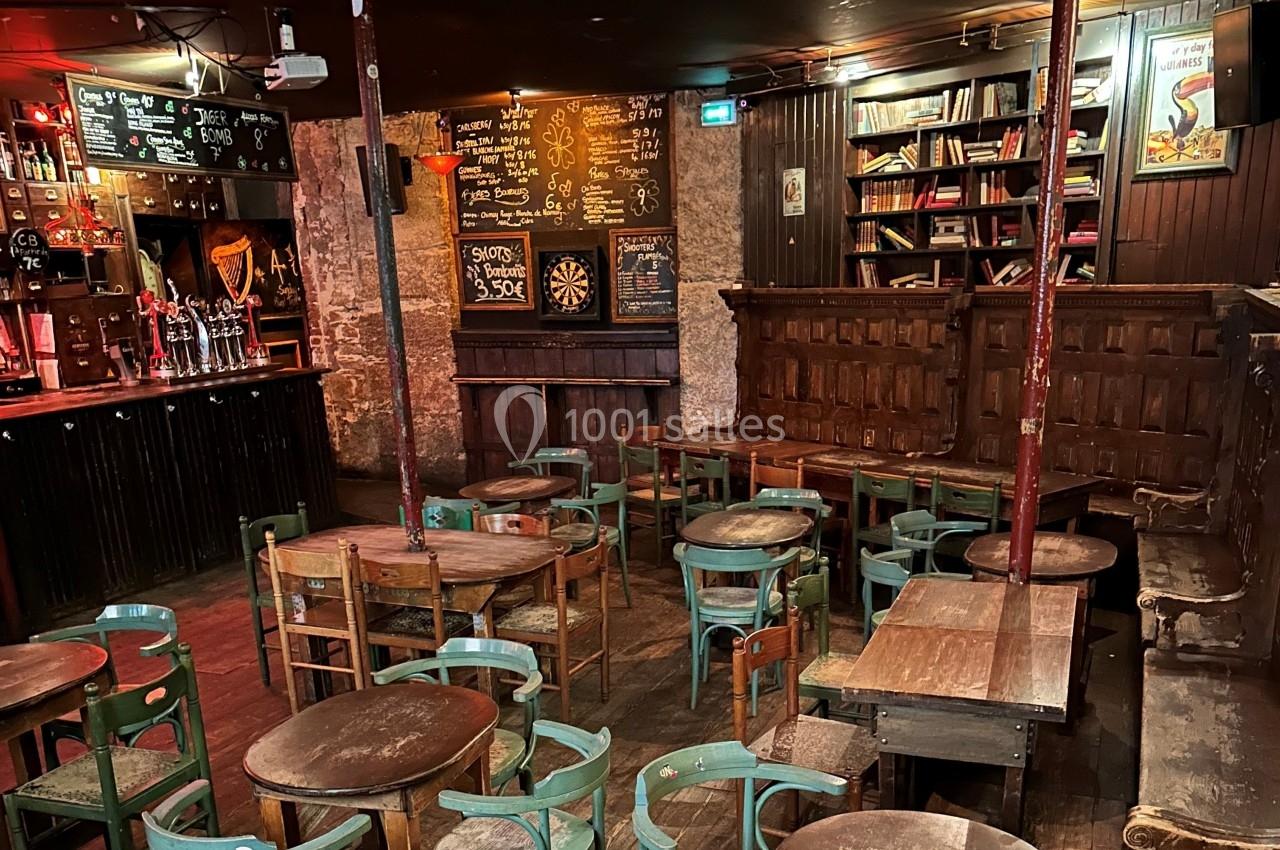 Salle de bar rustique avec tables en bois, chaises dépareillées, tableau noir, étagères de livres et ambiance chaleureuse.