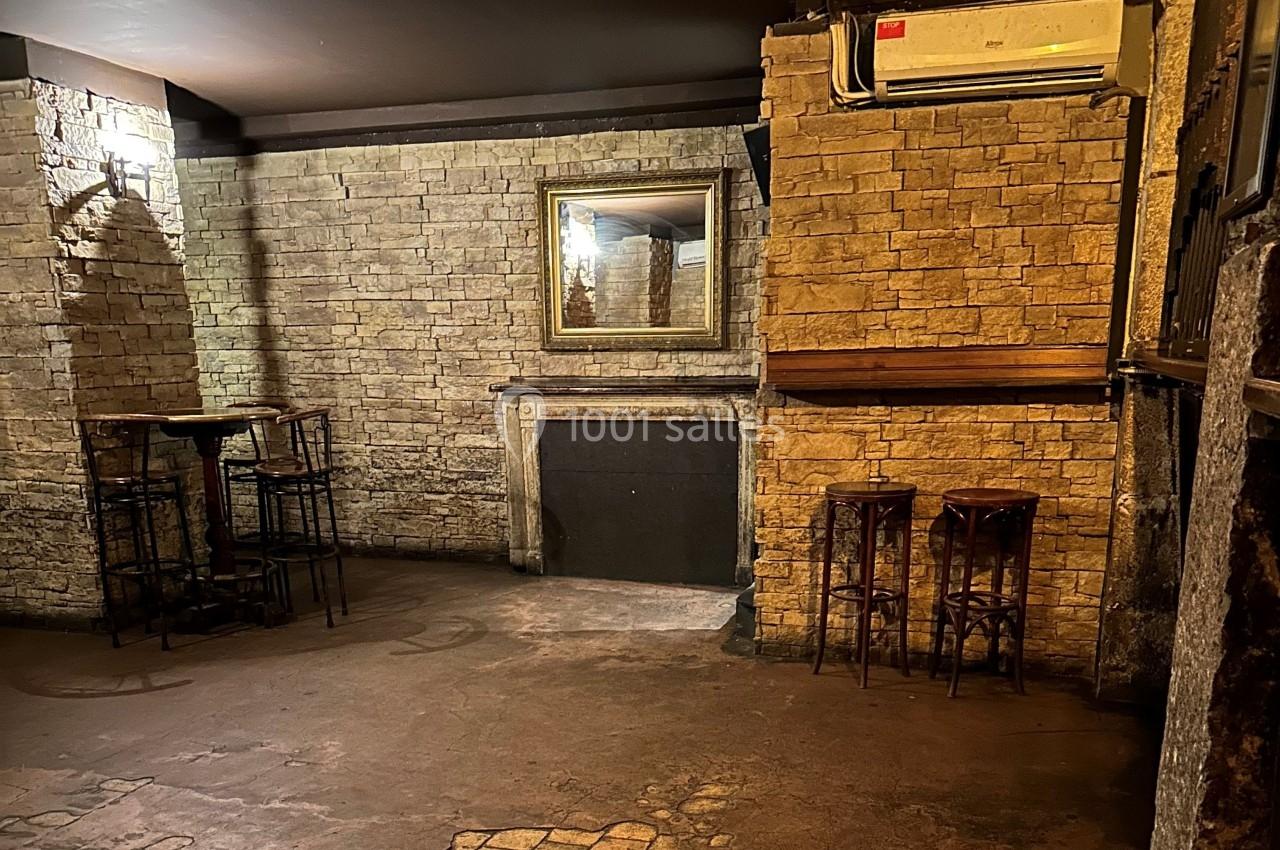 Salle intérieure avec murs en pierre, table haute avec tabourets à gauche, miroir et cheminée au centre.