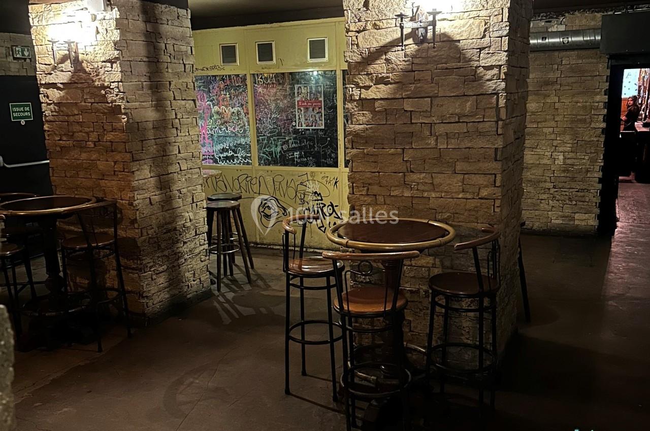 Salle avec murs en pierre, tables hautes en bois et tabourets, ambiance sombre avec graffitis sur une vitre au fond.