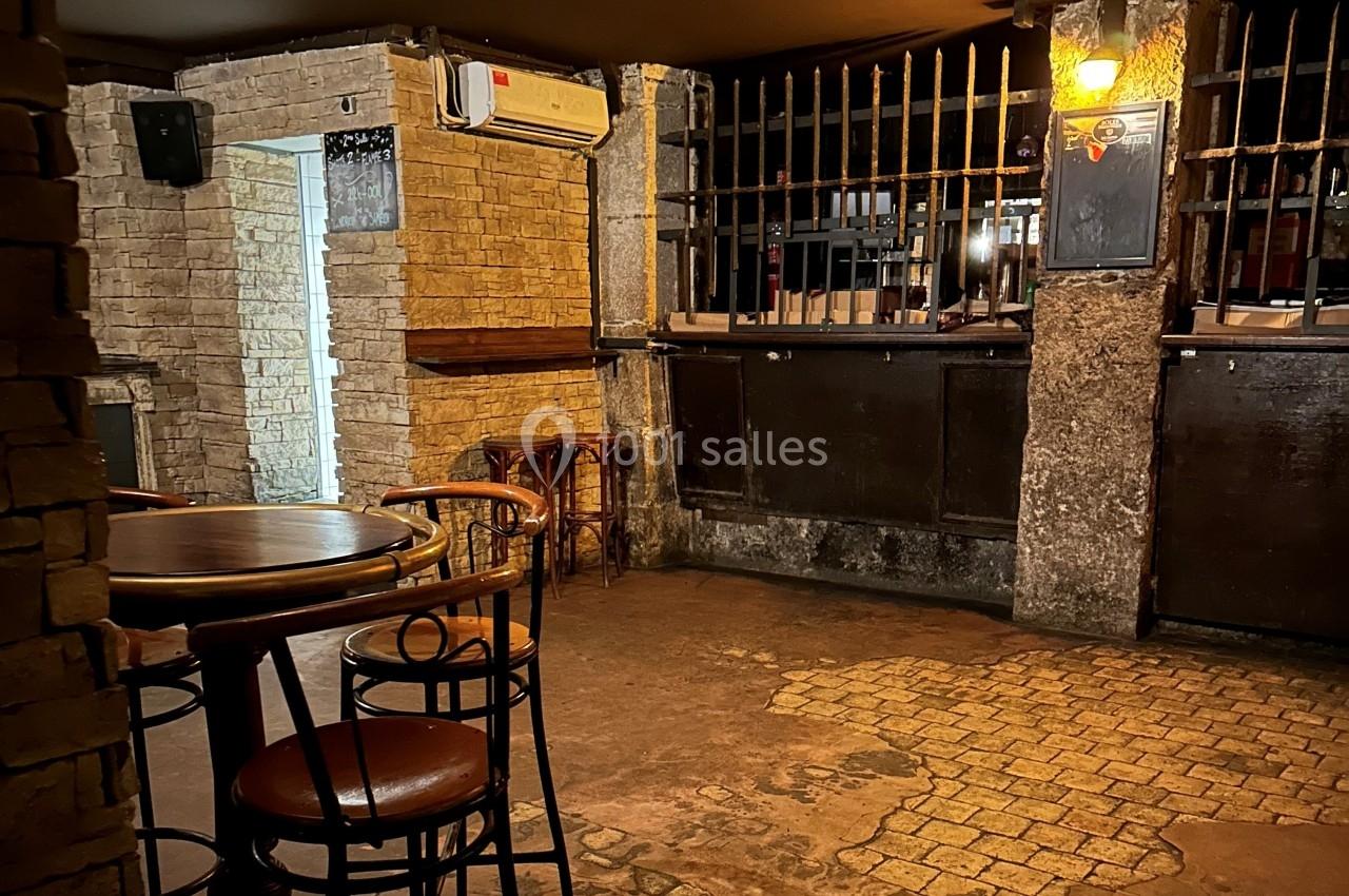 Intérieur d'un bar avec des tables en bois, des chaises hautes et un comptoir sombre derrière des barreaux métalliques.