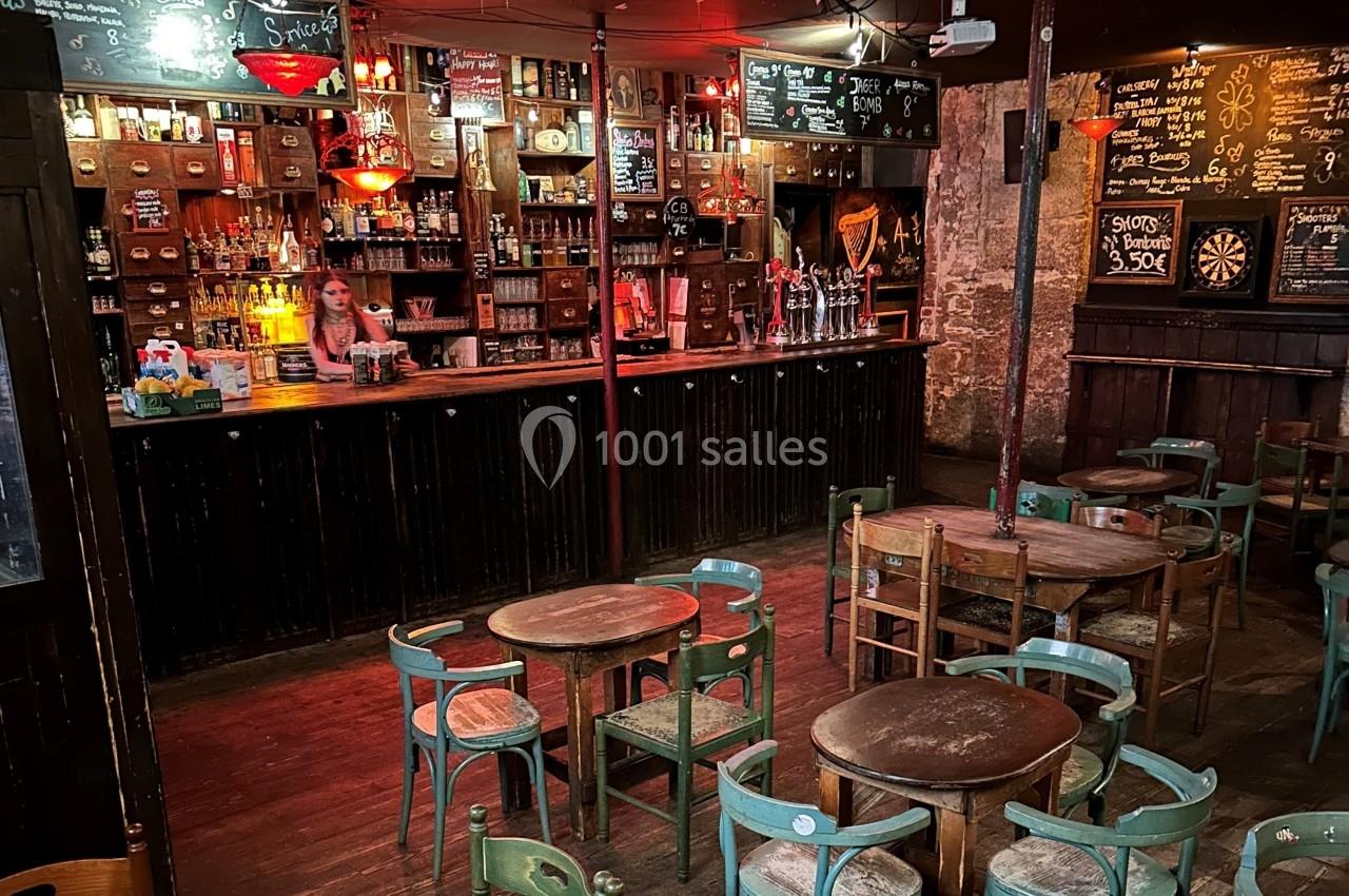 Intérieur d'un bar rustique avec tables en bois, chaises dispersées et une serveuse derrière le comptoir.