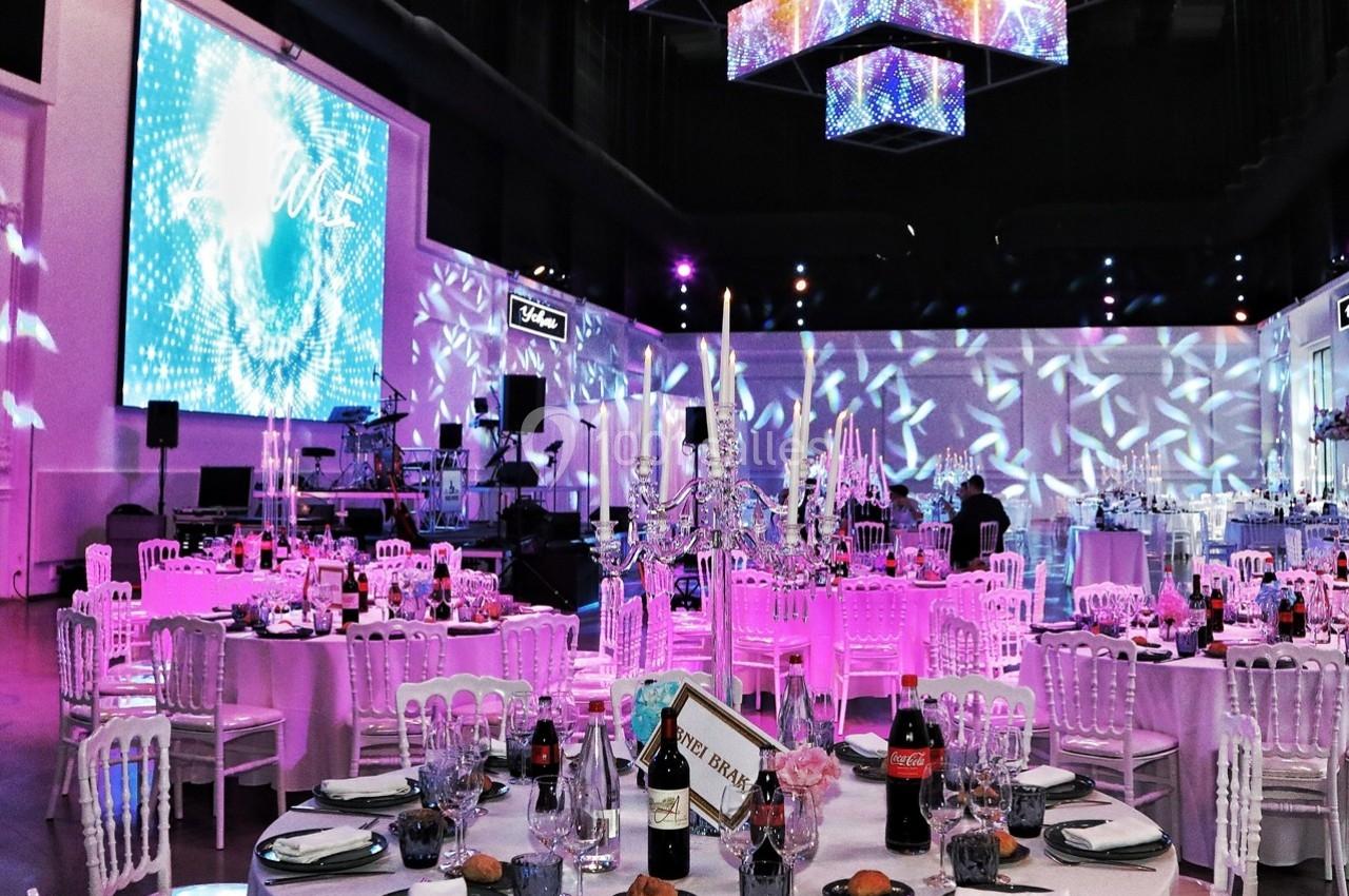 Salle de réception décorée avec des tables dressées, des chandeliers et des éclairages colorés suspendus.