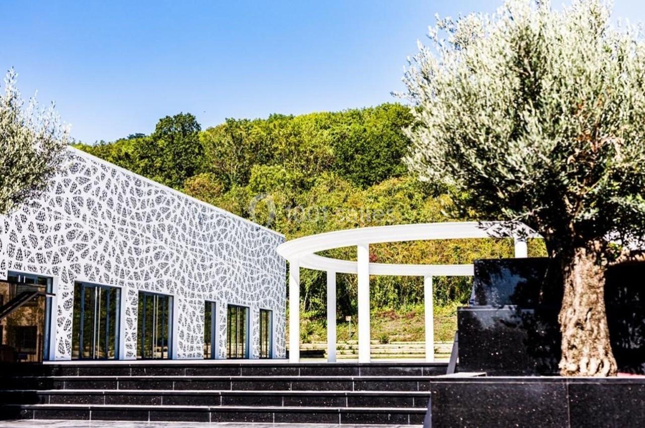 Bâtiment moderne avec façade blanche ajourée, entouré de verdure, escalier noir et olivier au premier plan.