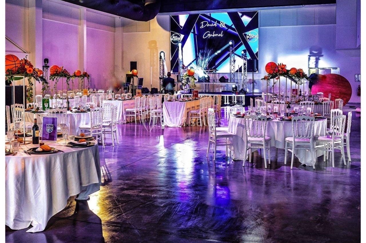 Salle de réception décorée avec des tables rondes dressées, des fleurs colorées et une scène avec écran lumineux en arrière…