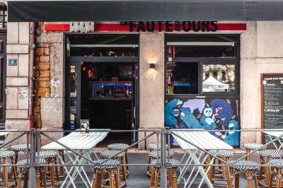 Terrasse de café avec tables et chaises alignées sur un trottoir, bordée de plantes et devantures de magasins.