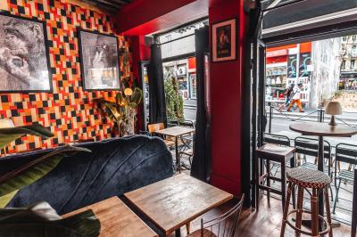 Terrasse de café avec tables et chaises alignées sur un trottoir, bordée de plantes et devantures de magasins.