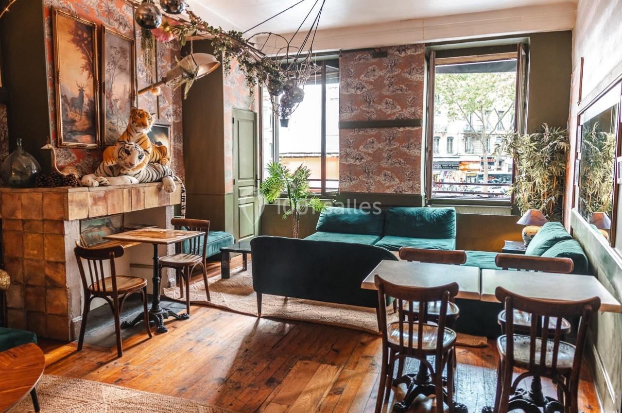 Salon lumineux avec mobilier vintage, canapé vert, tables en bois, cheminée décorative et vue sur la rue par la fenêtre.