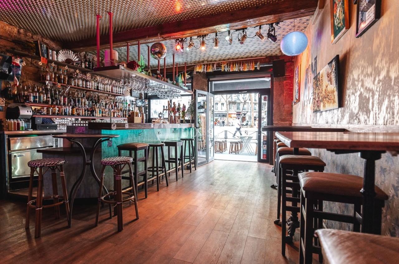 Intérieur d'un bar avec comptoir, tabourets, éclairage coloré et vue sur l'extérieur par une porte vitrée.