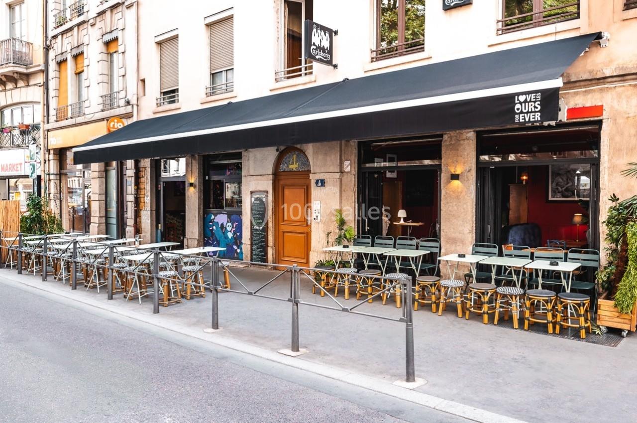 Terrasse de café avec tables et chaises alignées sous un grand auvent noir, située le long d'une rue urbaine.