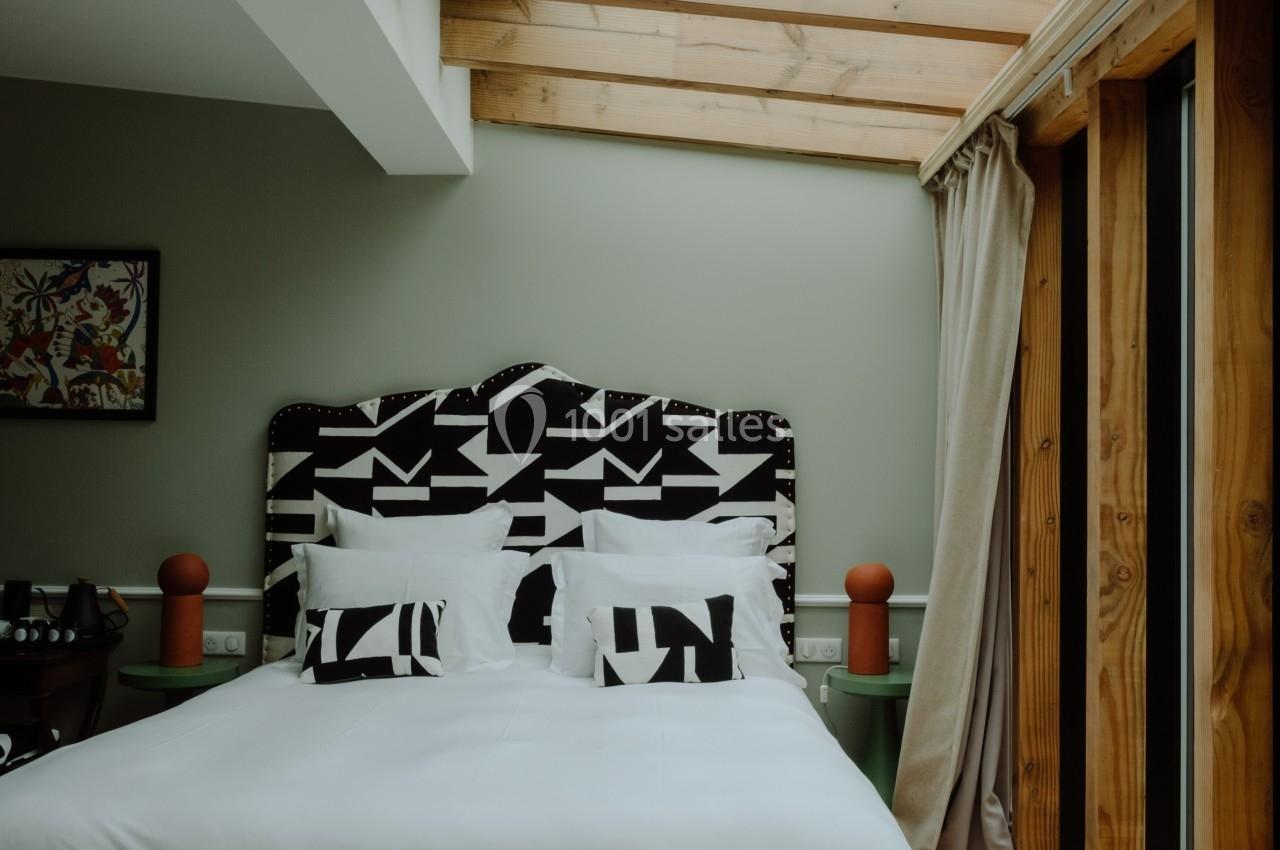 Chambre avec lit double, tête de lit à motifs géométriques, plafond en poutres apparentes et grande baie vitrée.