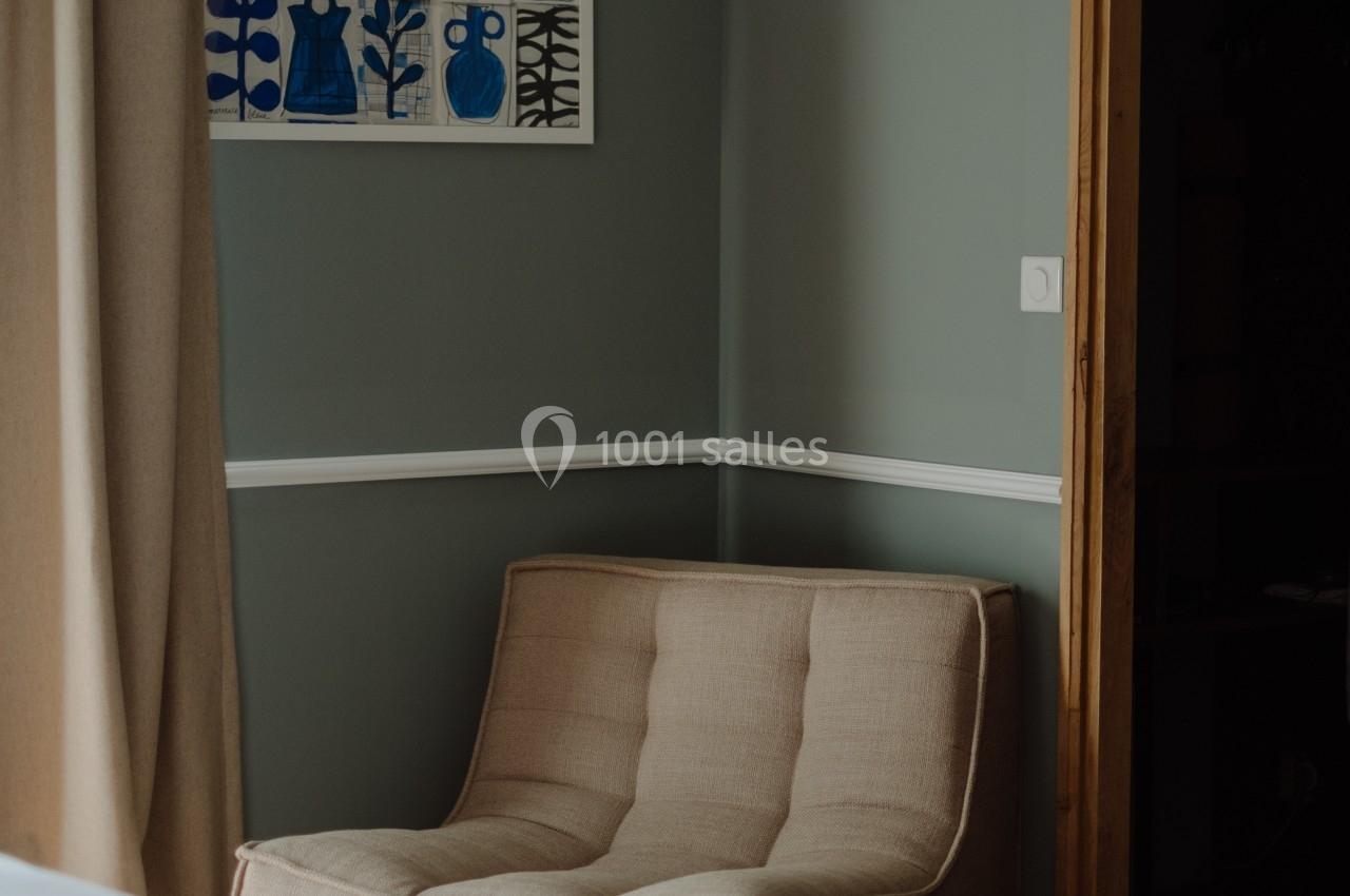 Fauteuil beige placé près d'un mur vert, avec un tableau encadré et des rideaux beiges en arrière-plan.