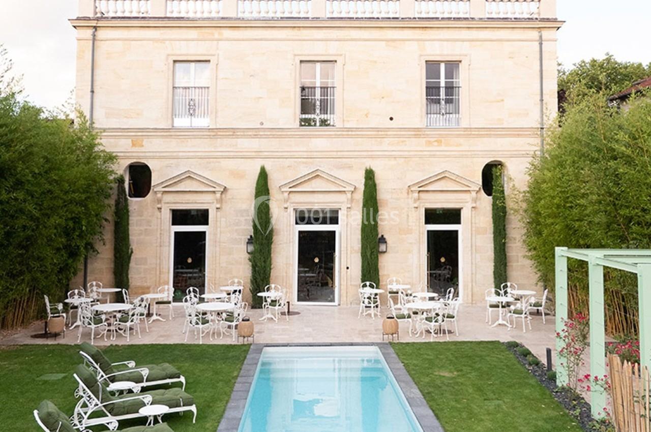 Façade d'un bâtiment en pierre avec terrasse aménagée, tables, chaises blanches et piscine entourée de verdure.