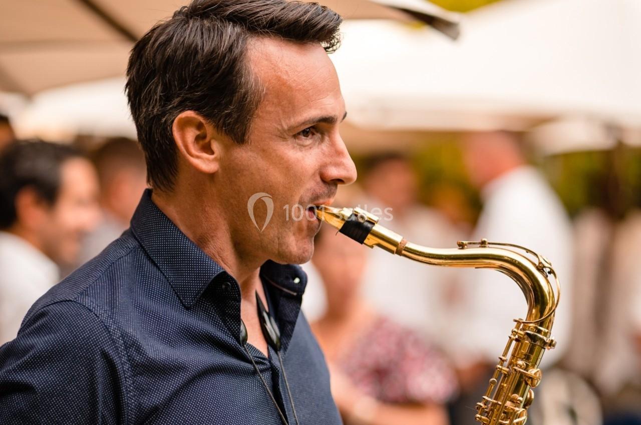 Un homme joue du saxophone dans un environnement extérieur avec des parasols et des personnes en arrière-plan flou.